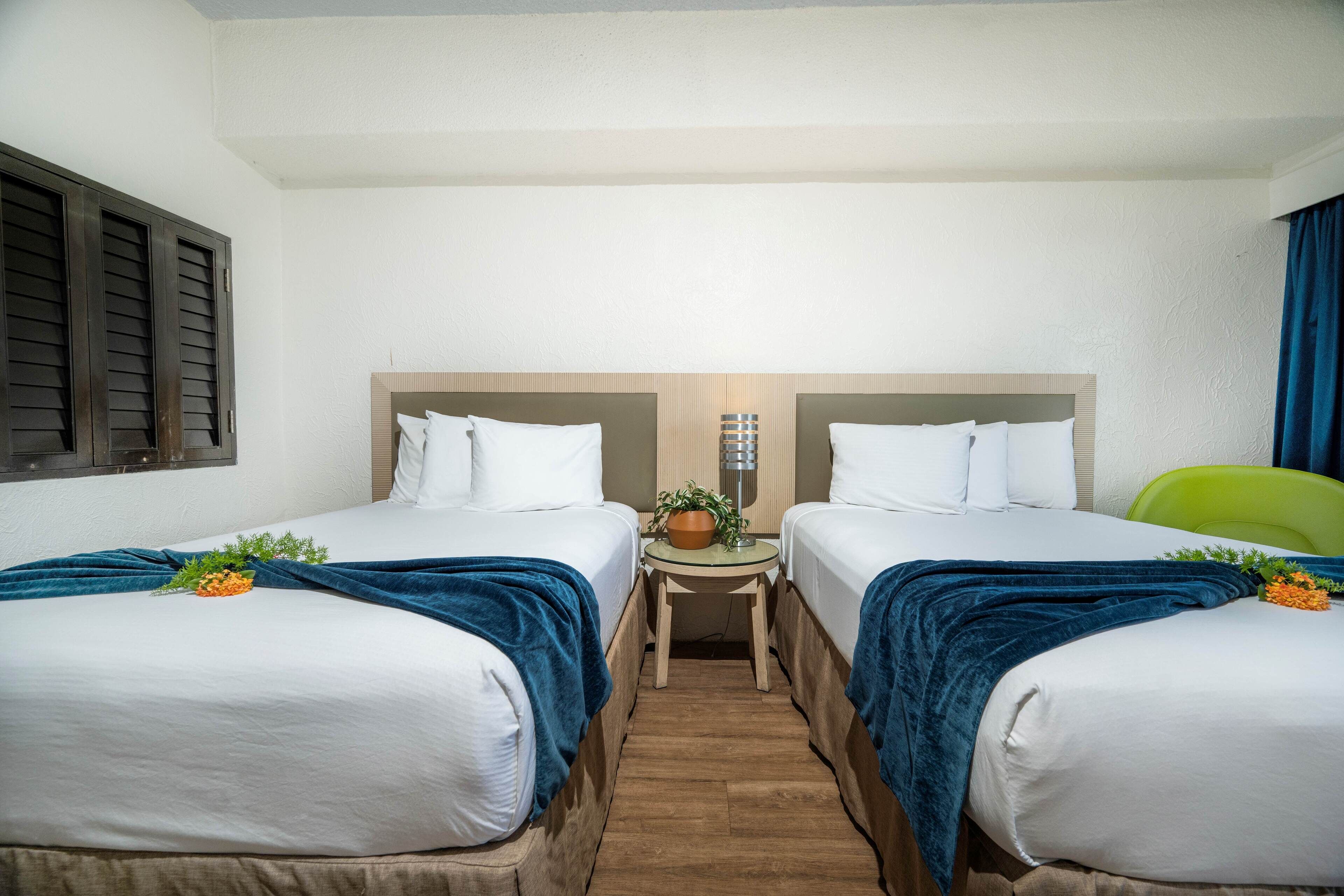 Quarto de hotel com duas camas, uma mesa pequena com abajur e um vaso de planta.