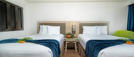 Quarto de hotel com duas camas, uma mesa pequena com abajur e um vaso de planta.