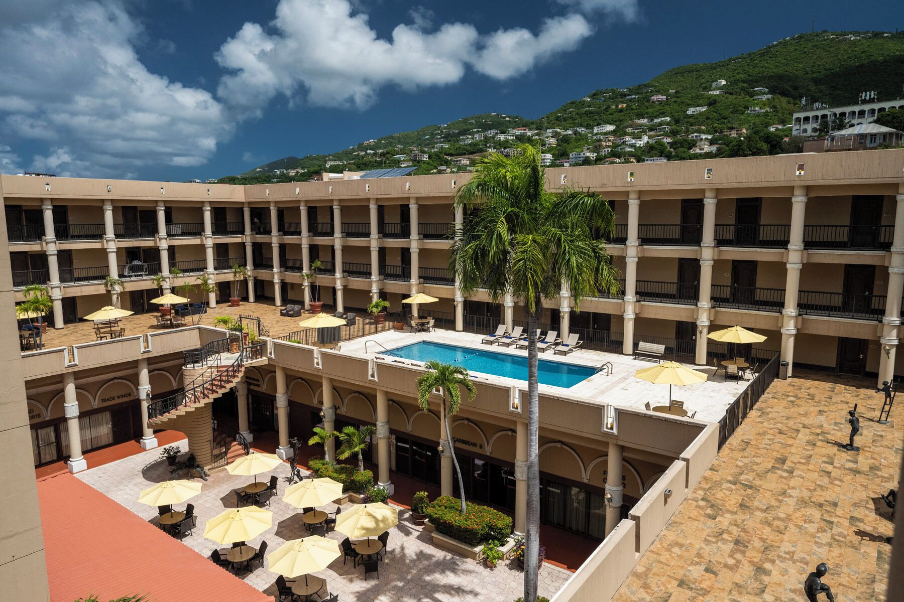 A multi-story hotel with a central swimming pool, outdoor seating areas, and a view of a hillside.