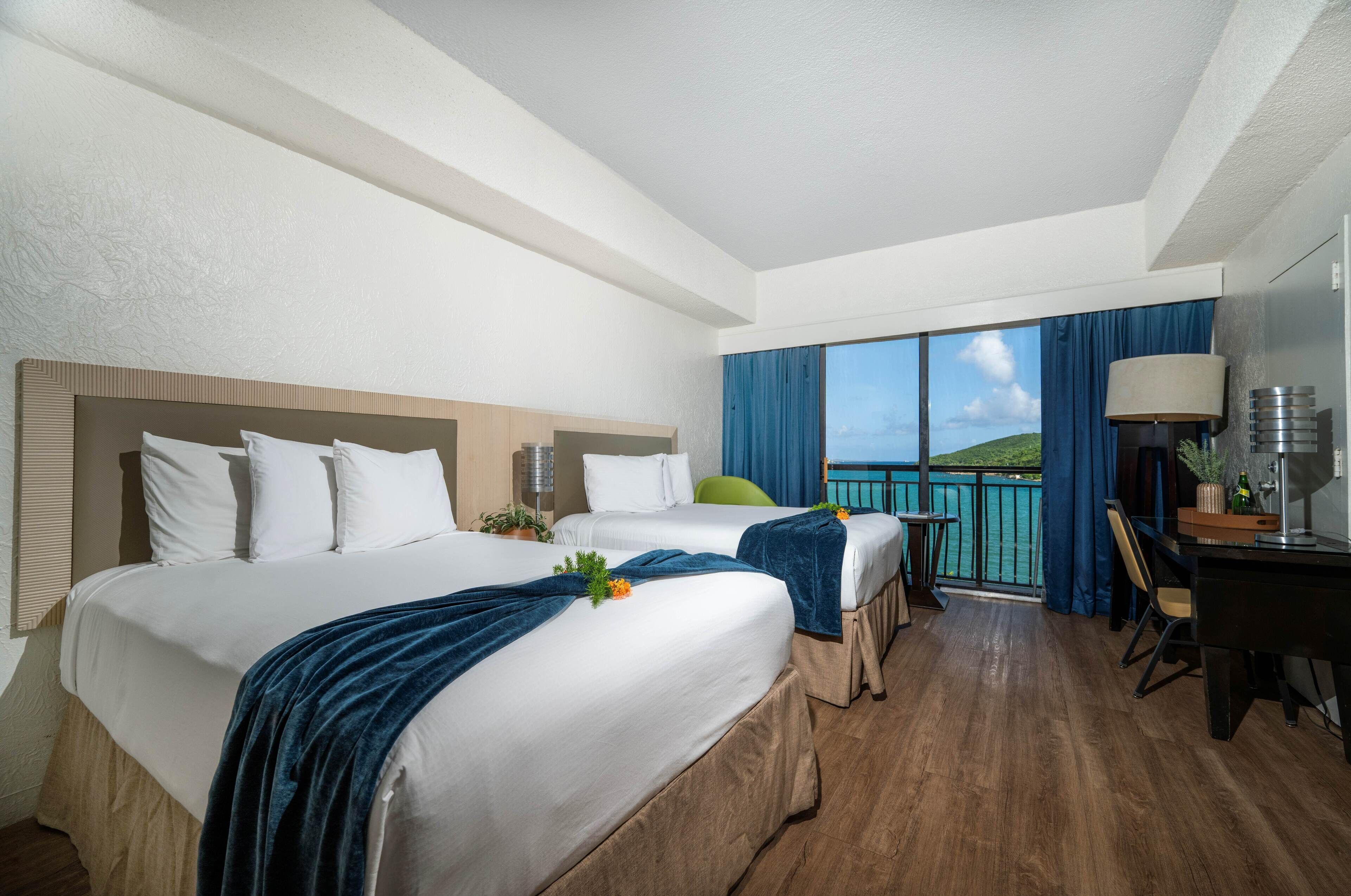 A hotel room with two beds, a balcony with a view, a desk, and a chair.