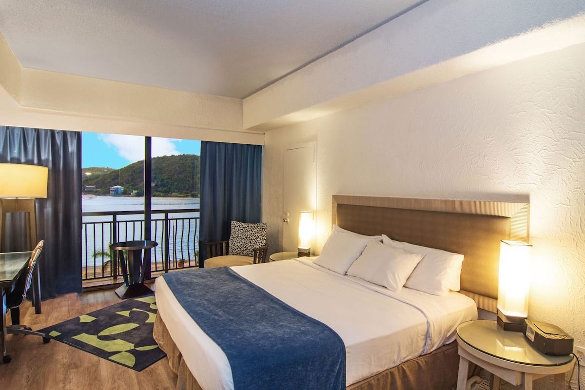 Ein Hotelzimmer mit Bett, Nachttischlampen, Schreibtisch, Stuhl, Blick auf ein Gewässer und einem Balkon.
