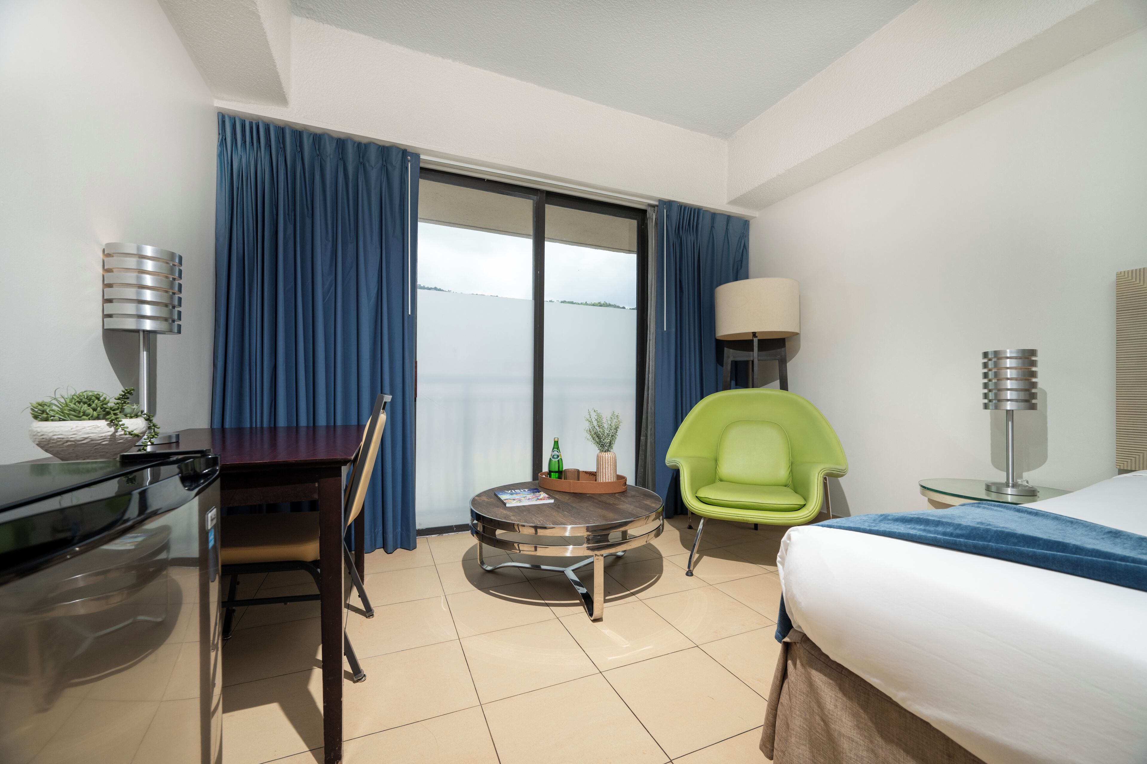 Ein Hotelzimmer mit Bett, einem runden Tisch mit Stühlen, einem Schreibtisch, einer Lampe und Meerblick durch das Fenster.