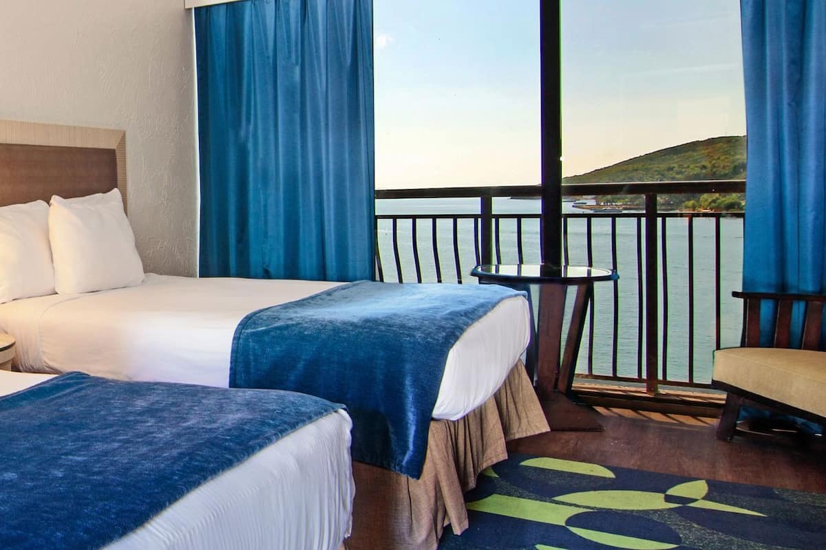 Ein Hotelzimmer mit zwei Betten, einem Nachttisch mit Lampe, einem kleinen Tisch und Blick aufs Meer.