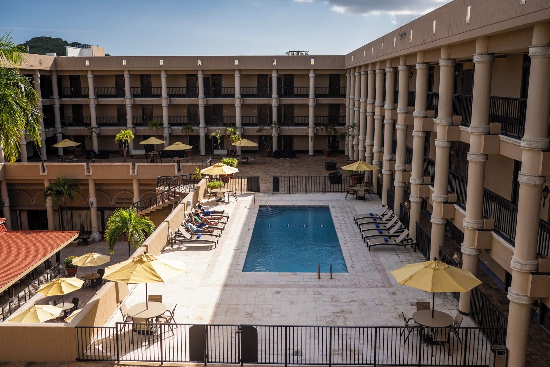 A hotel courtyard with a central pool, multiple balconies, outdoor seating, and palm trees.