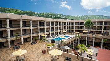 A multi-story hotel with a central courtyard featuring a swimming pool, outdoor seating, and a view of a green hillside.