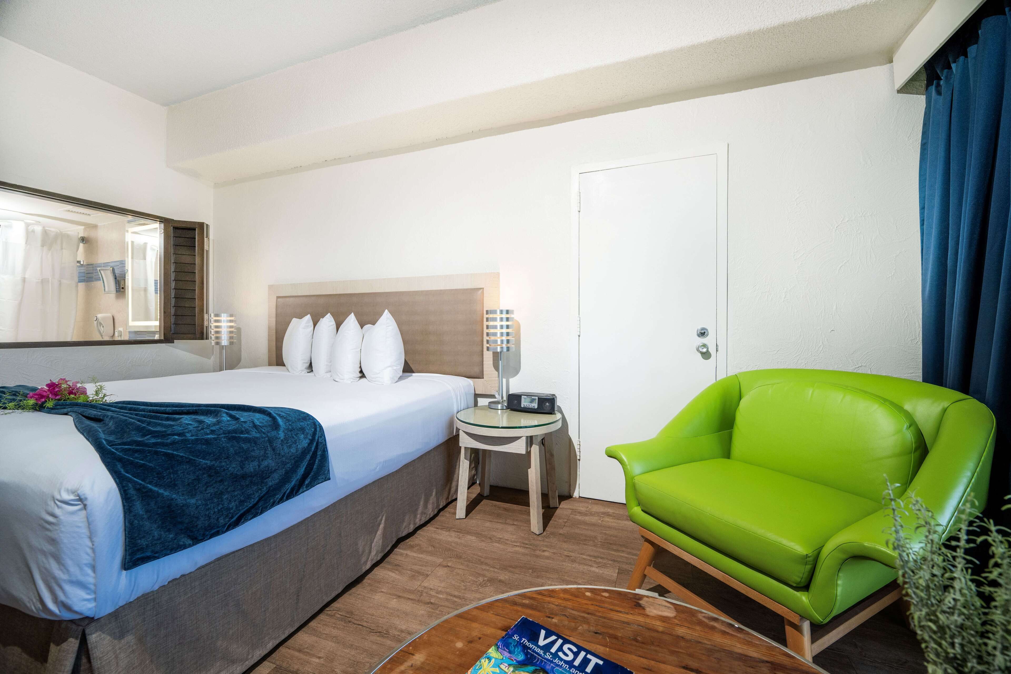Ein Hotelzimmer mit einem Bett, einem grünen Sessel, einem kleinen Tisch mit einer Zeitschrift und einer Tür.