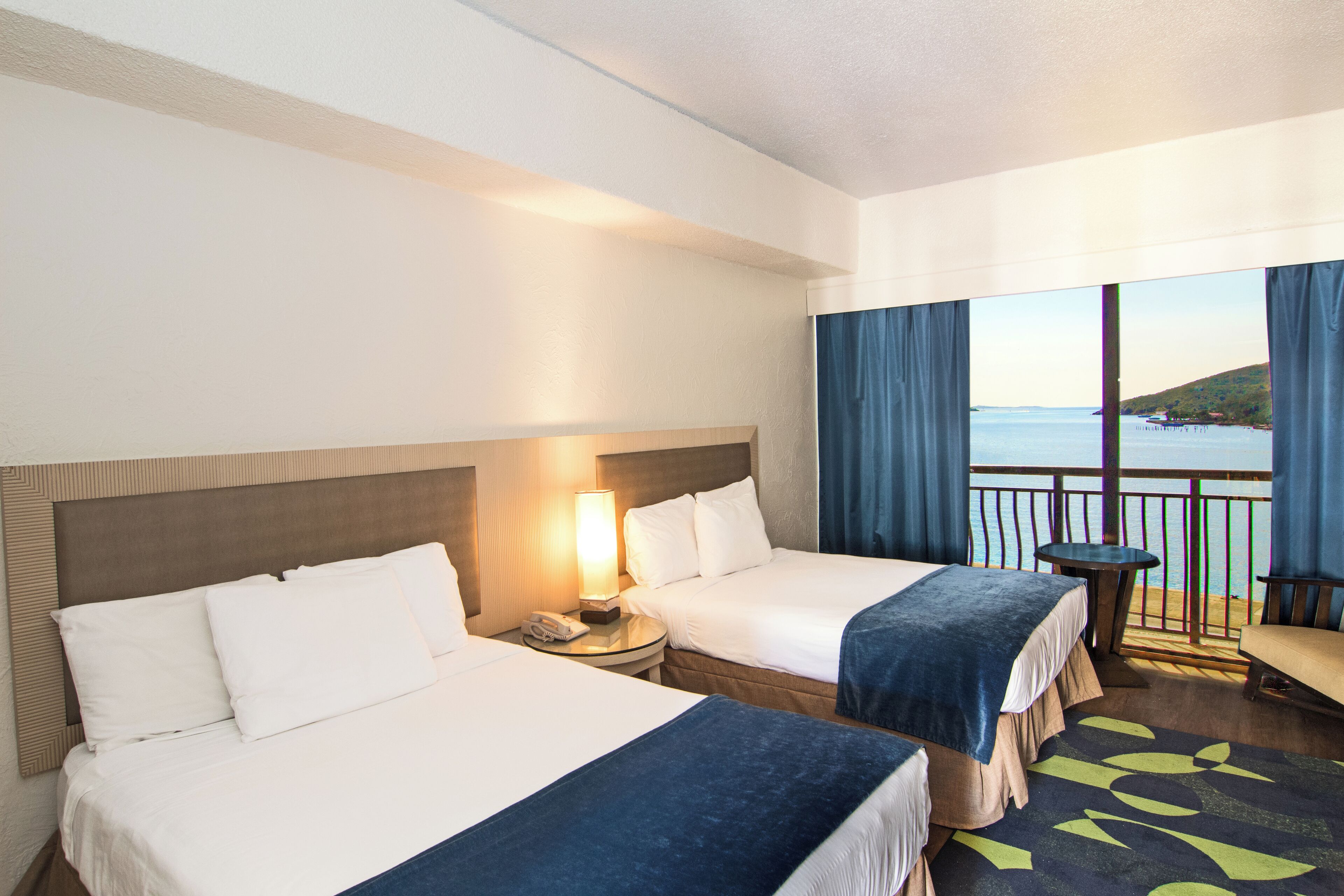 A hotel room with two beds, a nightstand with a lamp, a small table, a chair, and a balcony with a view of the sea.
