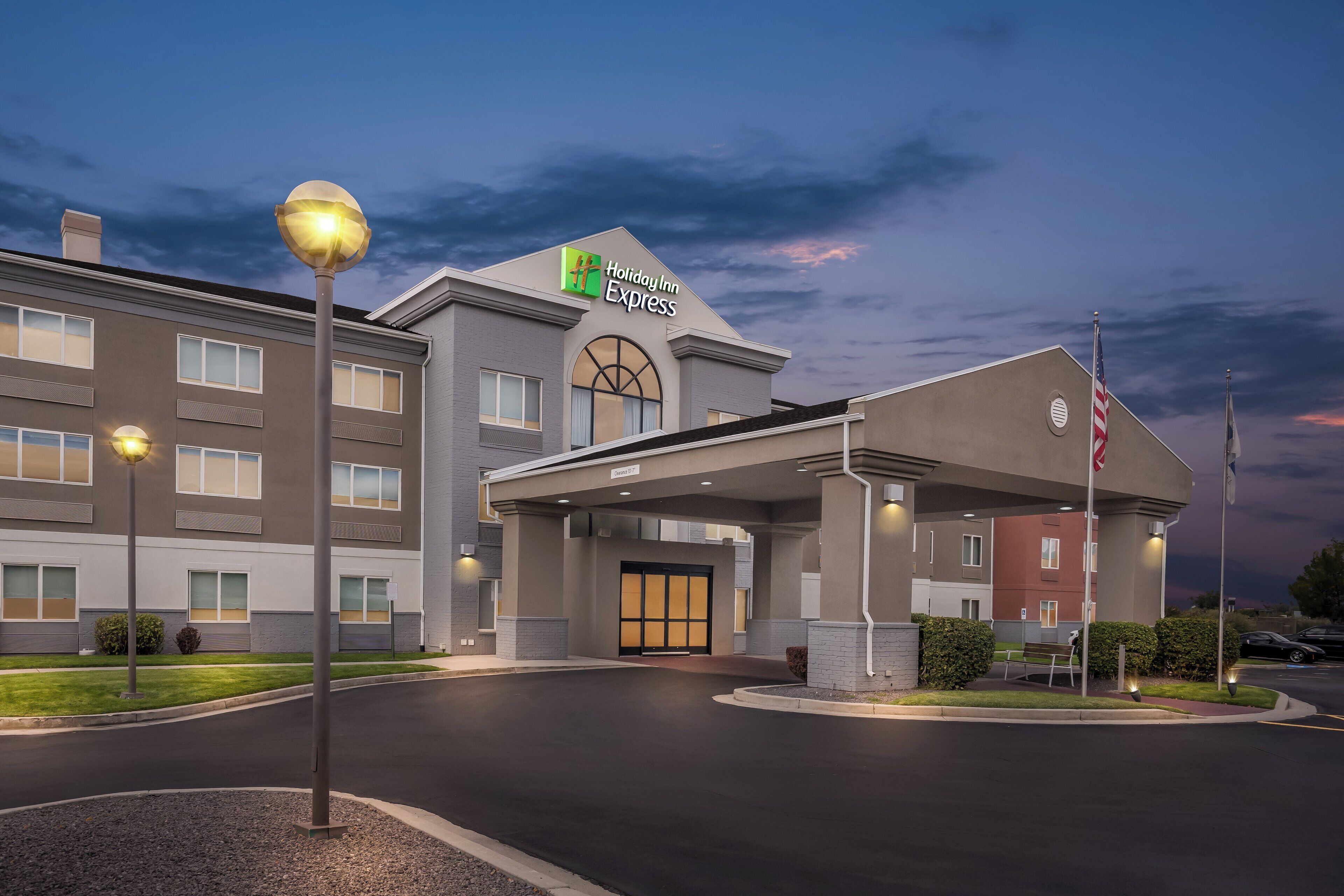 Edificio de Holiday Inn Express con una entrada bien iluminada, un área techada y una bandera estadounidense.