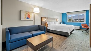 Une chambre d’hôtel avec un canapé bleu, une table basse en bois, un lit avec une literie blanche, une table de chevet et une lampe. Il y a une fenêtre donnant sur une vue sur les montagnes.