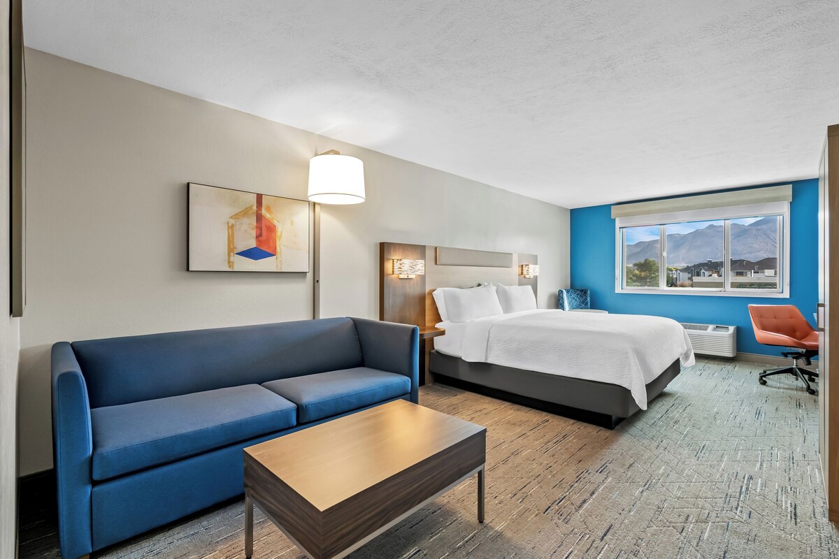 A hotel room with a blue sofa, a wooden coffee table, a bed with white bedding, a nightstand, and a lamp. There is a window with a mountain view.