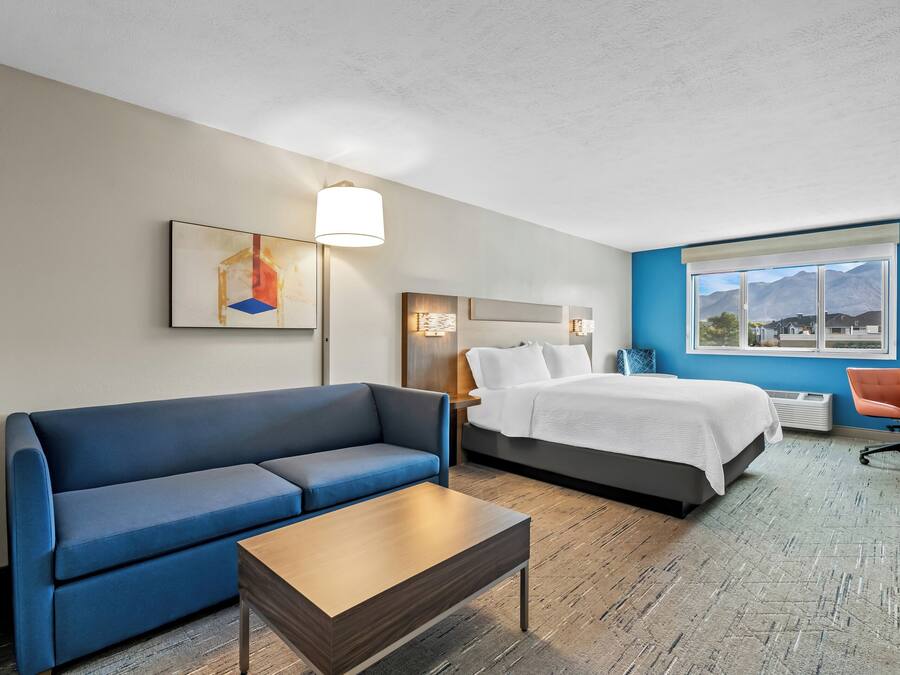 Une chambre d’hôtel avec un canapé bleu, une table basse en bois, un lit avec une literie blanche, une table de chevet et une lampe. Il y a une fenêtre donnant sur une vue sur les montagnes.