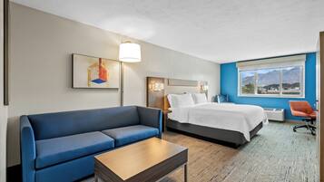Quarto de hotel moderno com sofá azul, mesa de centro de madeira, cama com roupa de cama branca, mesa de cabeceira, luminária e vista para montanhas pela janela.