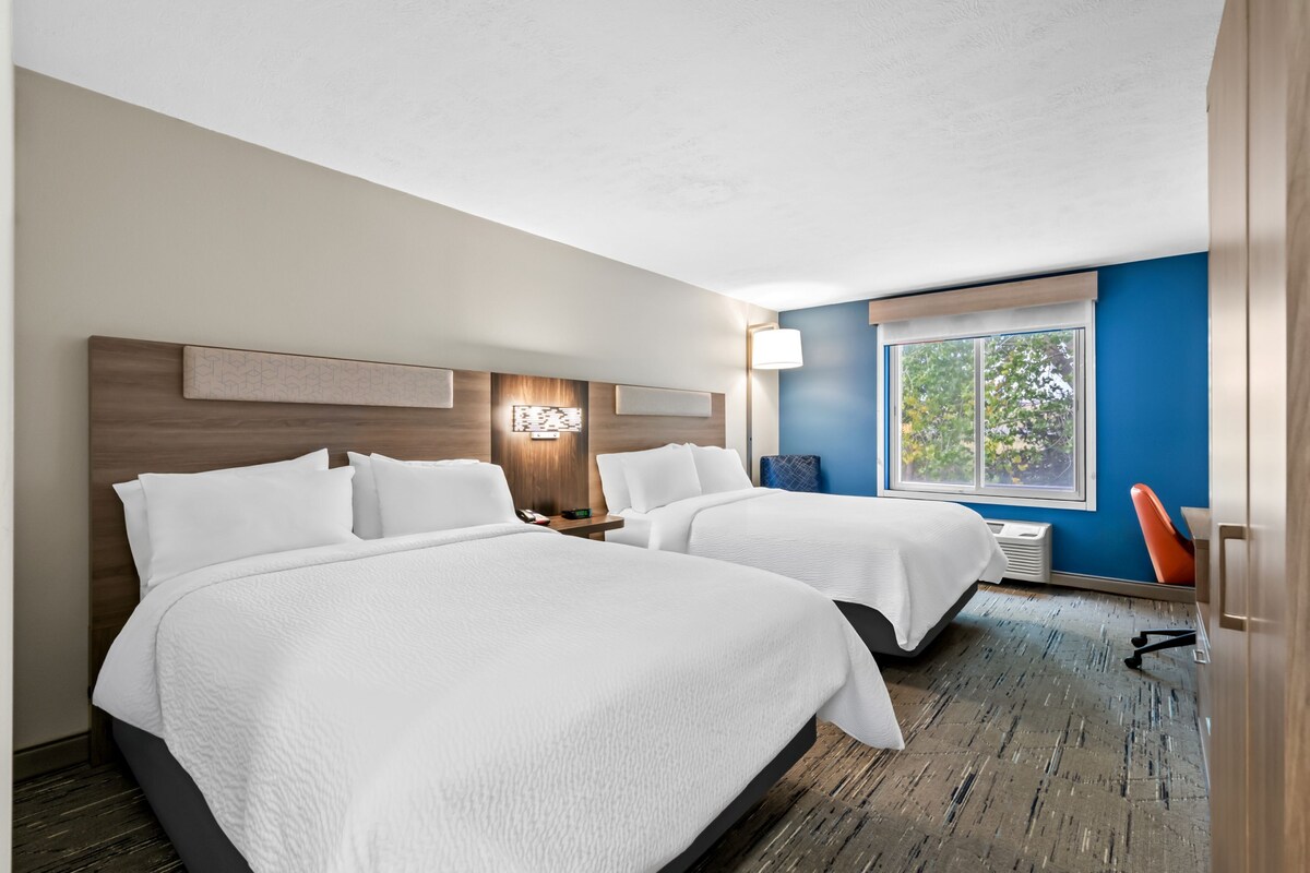 A hotel room with two beds, a nightstand, a lamp, and a window with a view of trees.