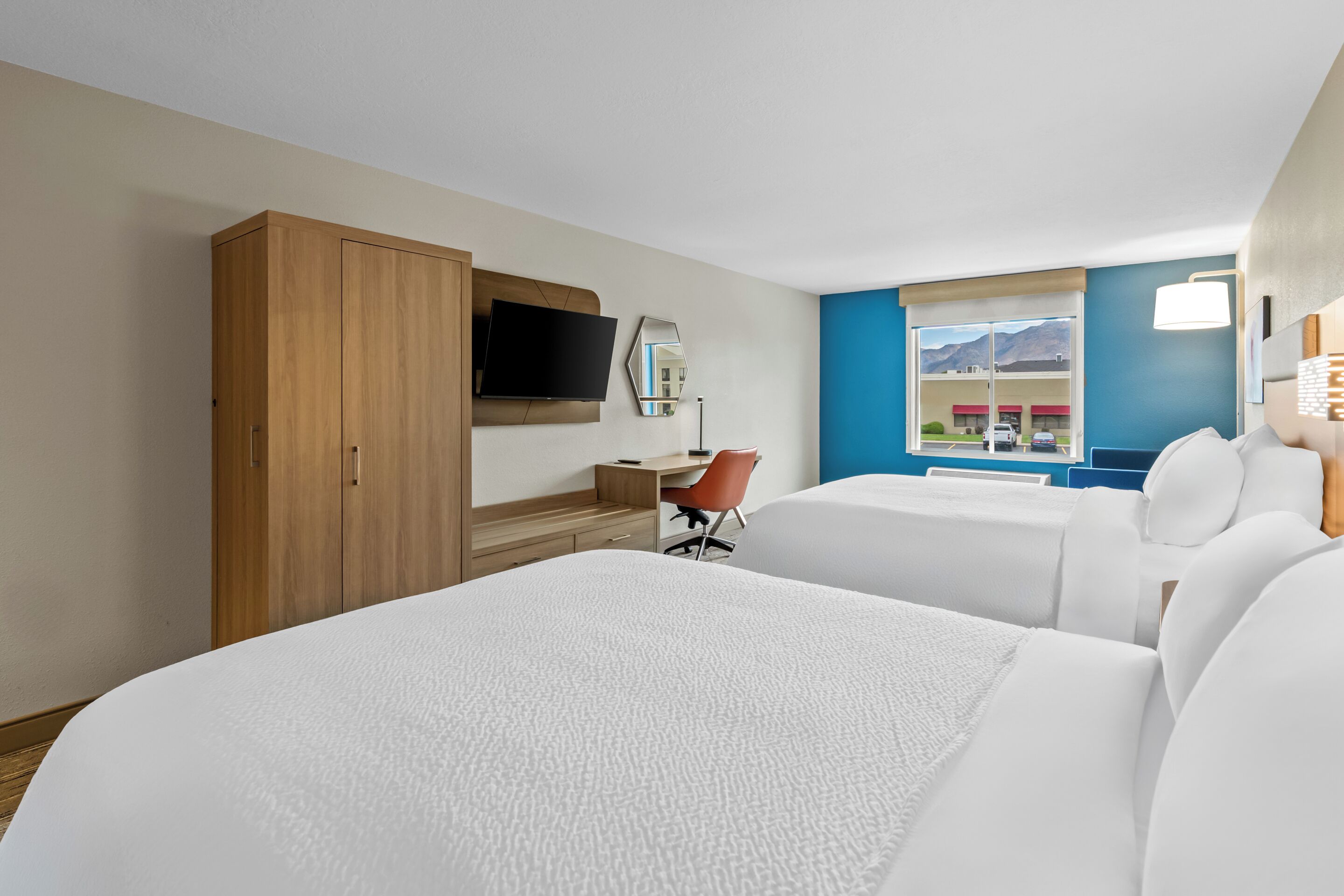 A hotel room with two beds, a desk, a chair, a TV, and a window with a mountain view.