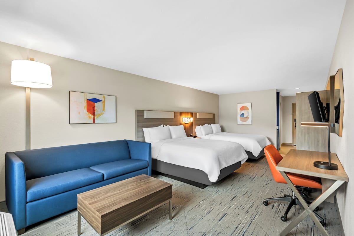 A hotel room with a blue sofa, two beds, a desk with a chair, and a lamp.