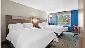 Une chambre d’hôtel avec deux lits, une table de chevet, une lampe, une fenêtre donnant sur la verdure et un mur d’accent bleu.