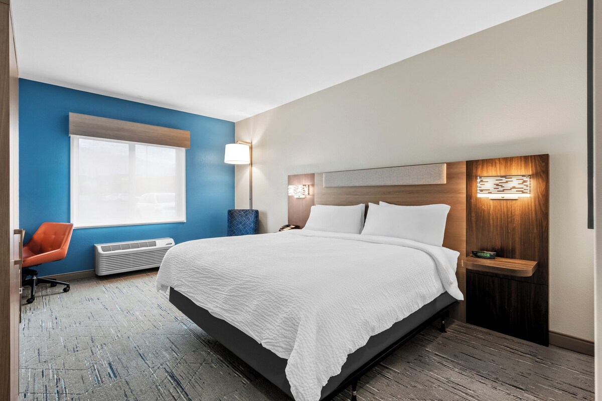 A hotel room with a large bed, a desk, a chair, a window, and a wall-mounted light fixture.
