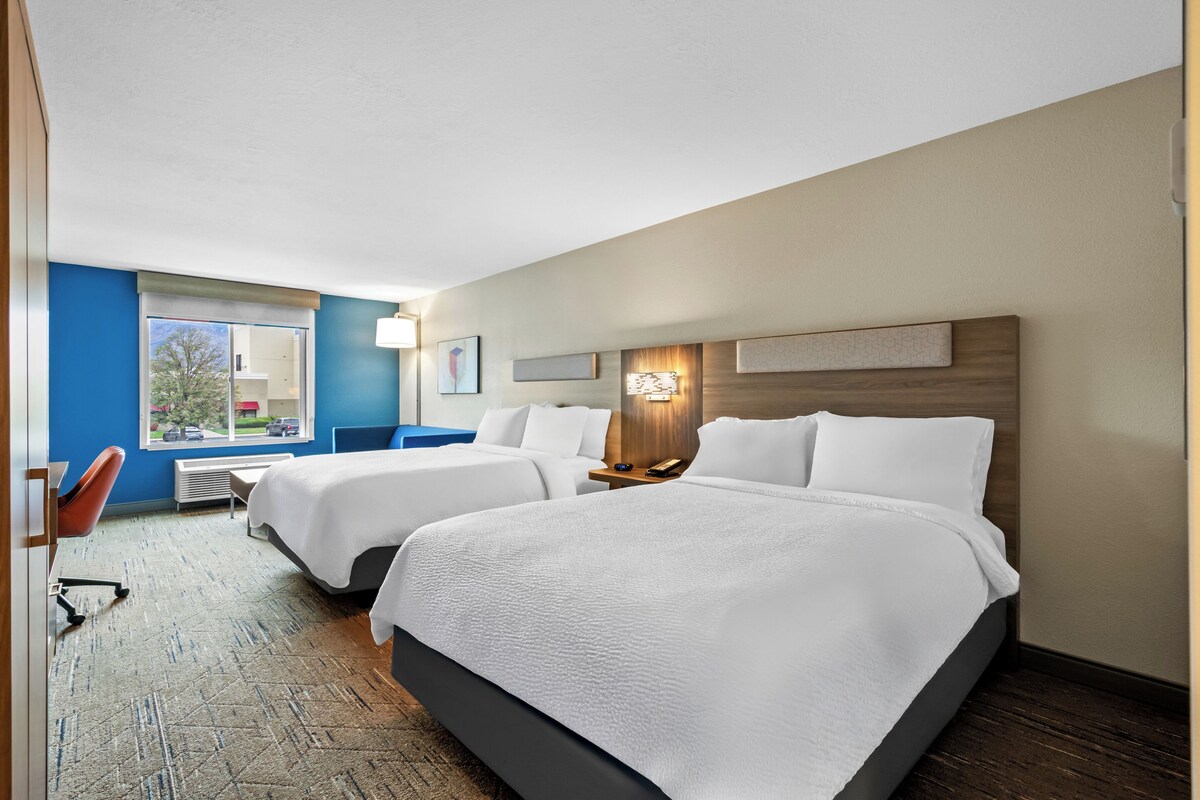 A hotel room with two beds, a desk, a chair, and a window with a view of trees.
