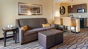 A hotel room with a sofa, ottoman, a small table with a phone, a framed picture on the wall, a kitchen area with a microwave, and a bar area with chairs. - Comfort Inn & Suites Market - Airport (Great Falls)