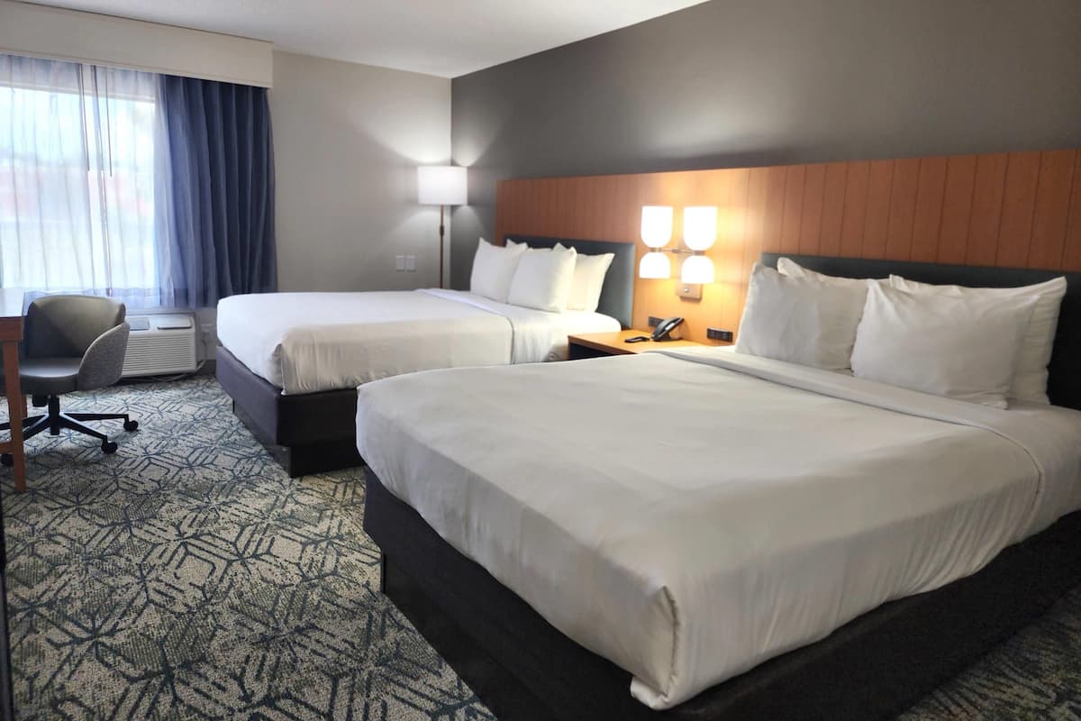 A hotel room with two beds, a desk, and a chair. There is a lamp on the desk and a phone on the nightstand.