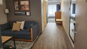 A hotel room with a sofa, a small table, a wooden bench, a bed, and a hallway leading to another room. - Country Inn & Suites by Radisson, Dahlgren-King George, VA (King George)