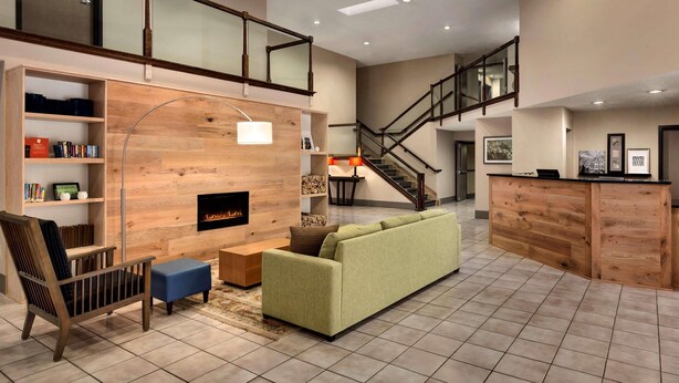 A spacious living area with a high ceiling, a fireplace, a green sofa, a wooden bookshelf, and a staircase leading to an upper level. - Country Inn & Suites by Radisson, Dahlgren-King George, VA (King George)