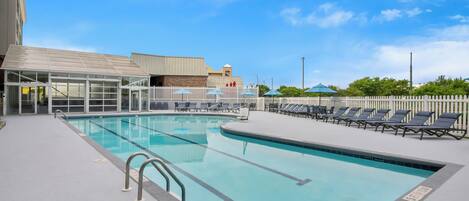 A swimming pool with lounge chairs and umbrellas, surrounded by a white fence and a building with a glass facade.