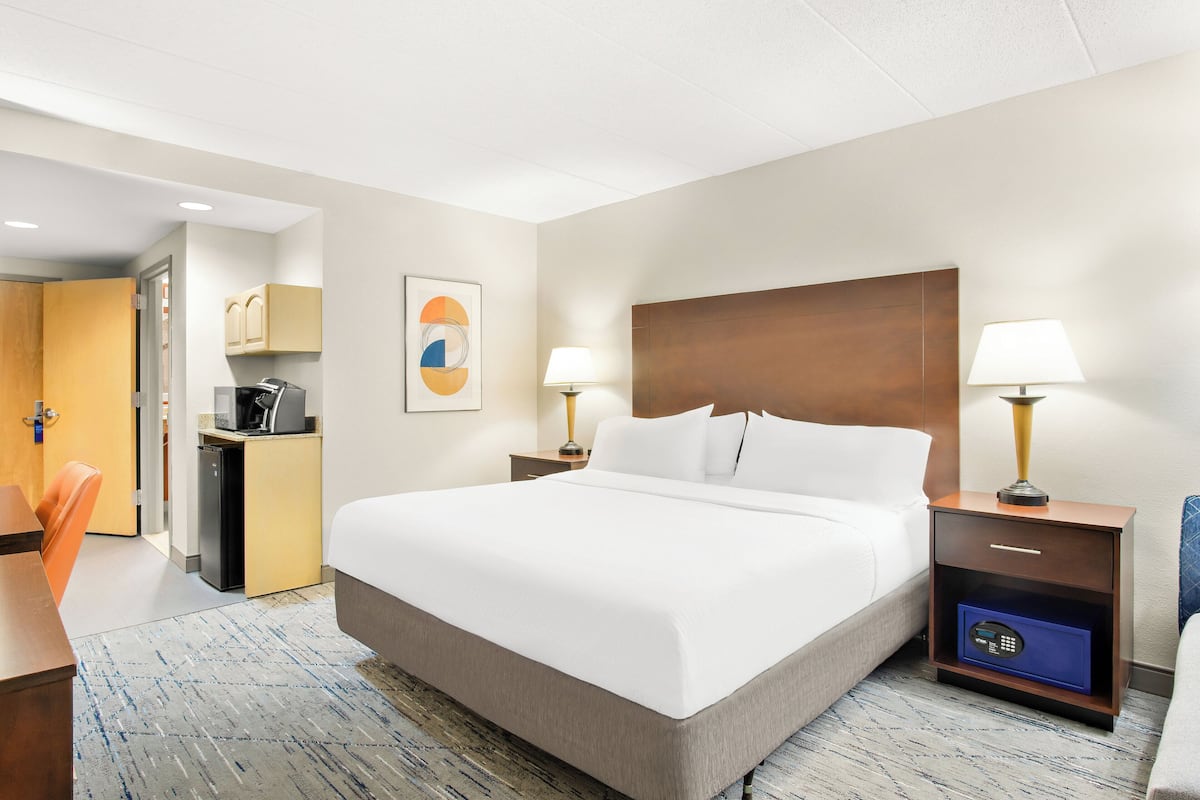 A hotel room with a bed, bedside tables with lamps, a desk with a computer, and a small kitchen area.