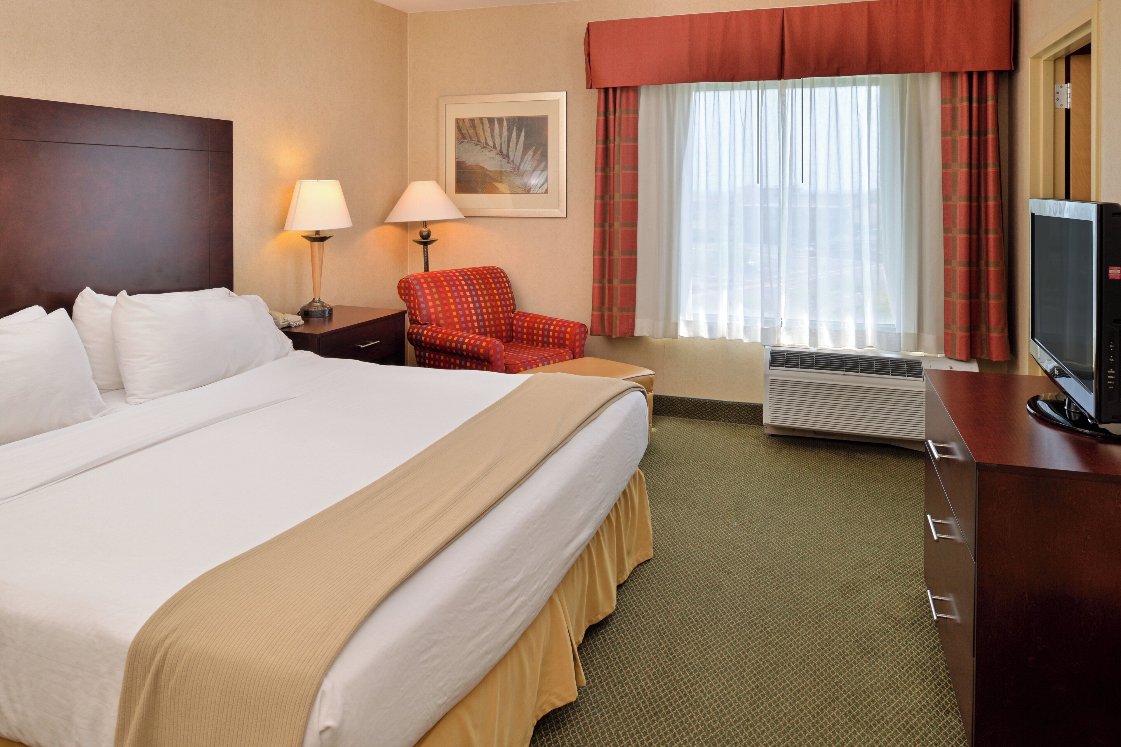 Habitación de hotel con una cama grande, un escritorio con televisor, una silla, una ventana con cortinas y un aire acondicionado instalado en la pared.