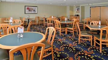 A hotel dining area with round tables and wooden chairs, carpeted floor, and framed artwork on the walls.
