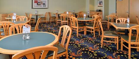 A hotel dining area with round tables and wooden chairs, carpeted floor, and framed artwork on the walls.