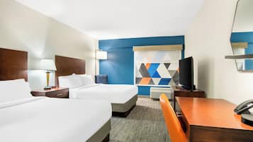 Quarto de hotel com duas camas, uma escrivaninha, uma cadeira, uma televisão e um detalhe decorativo na parede.