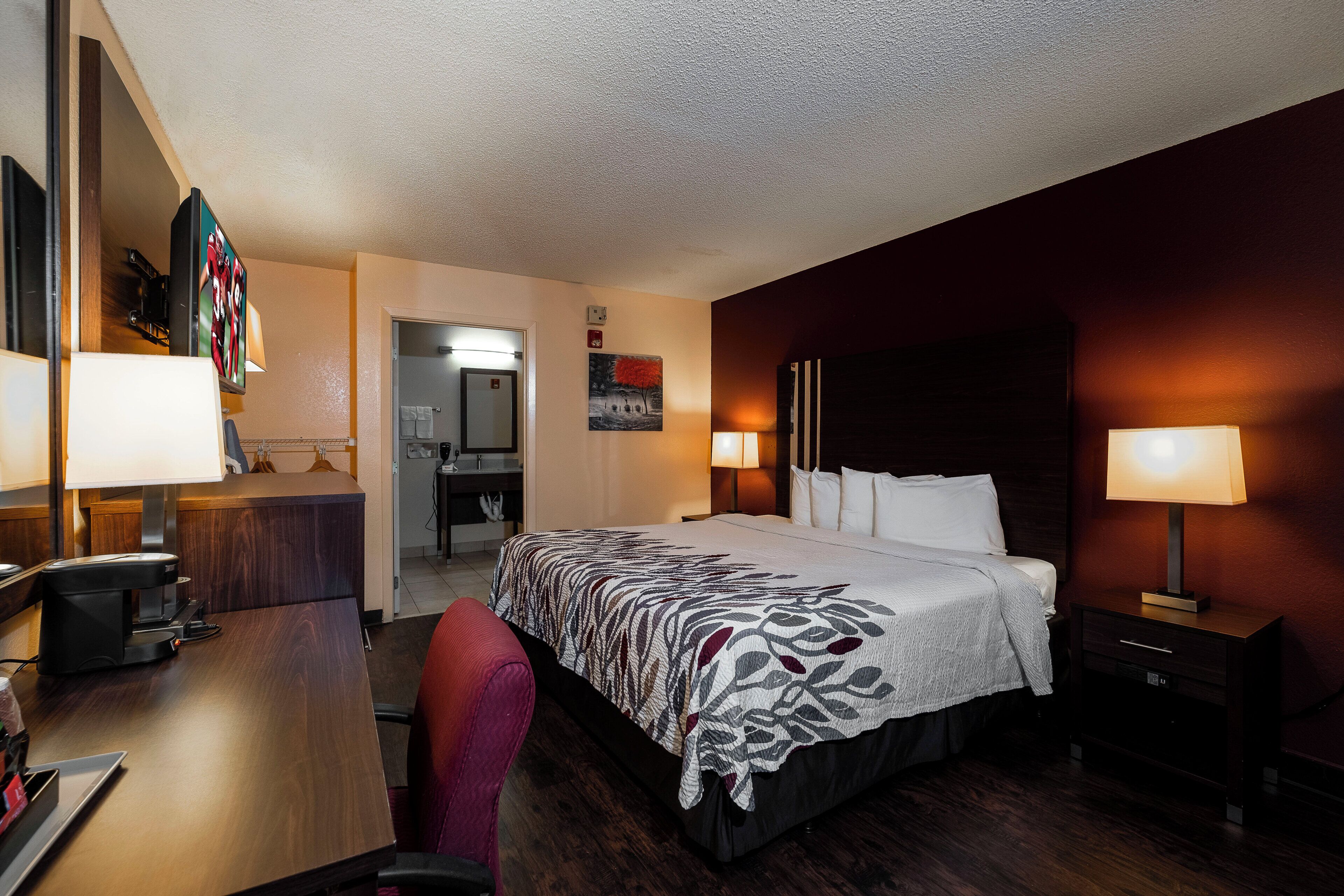 A hotel room with a bed, bedside lamps, a desk with a coffee maker, a chair, and a painting on the wall.