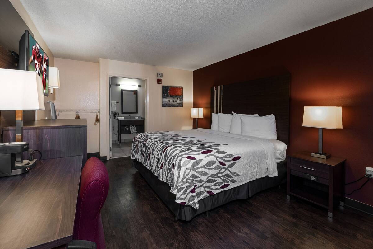 A hotel room with a bed, bedside table, desk, chair, and a painting on the wall.