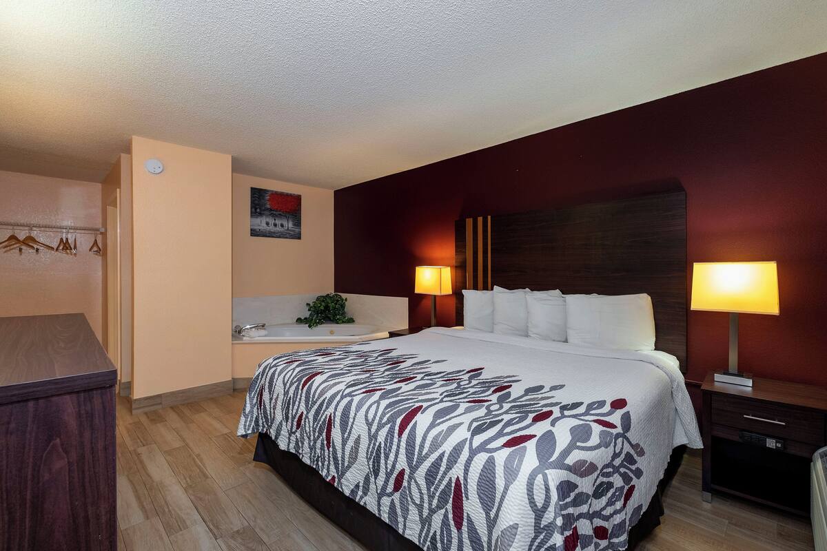 Ein Hotelzimmer mit Bett, Nachttischlampen, einem Nachttisch, einem Schminktisch, einem Badezimmer mit Waschbecken und Spiegel sowie einem Wandhandtuchhalter.