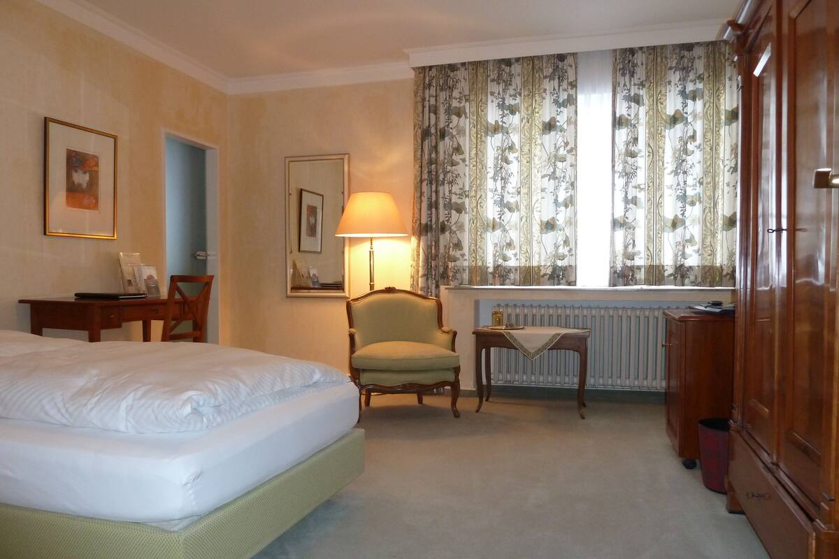 Una habitación de hotel con una cama, una silla, un escritorio, una lámpara y un armario.