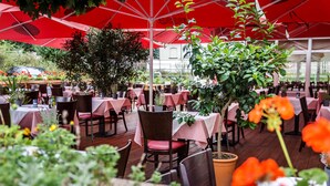An outdoor dining area with tables and chairs under red umbrellas, surrounded by potted plants and greenery. - AKZENT Hotel Frankenberg (Frankenberg)