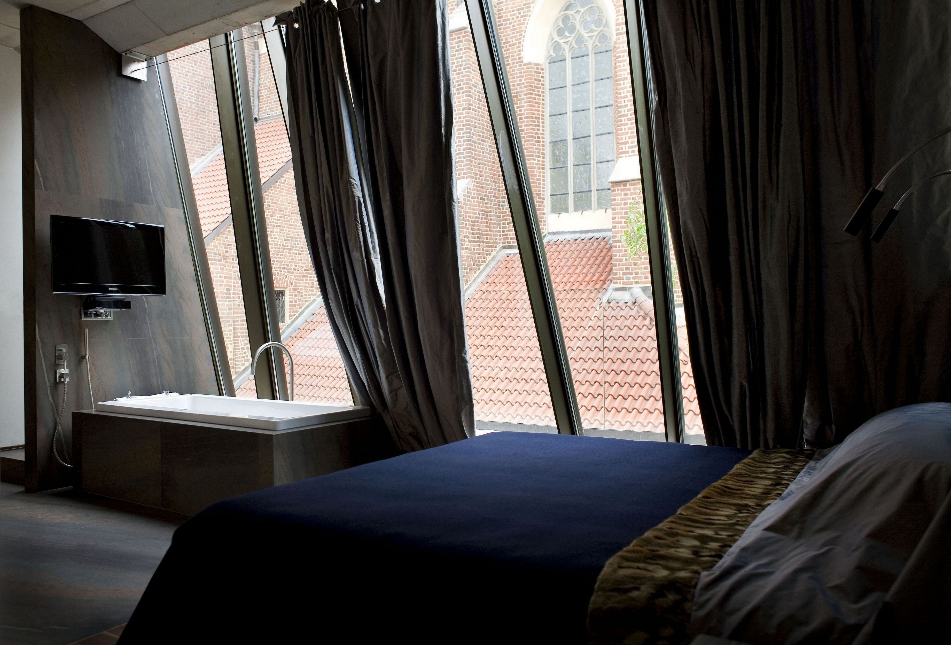 A hotel room with a large bed, a TV mounted on the wall, a window with curtains, and a view of a brick building outside.