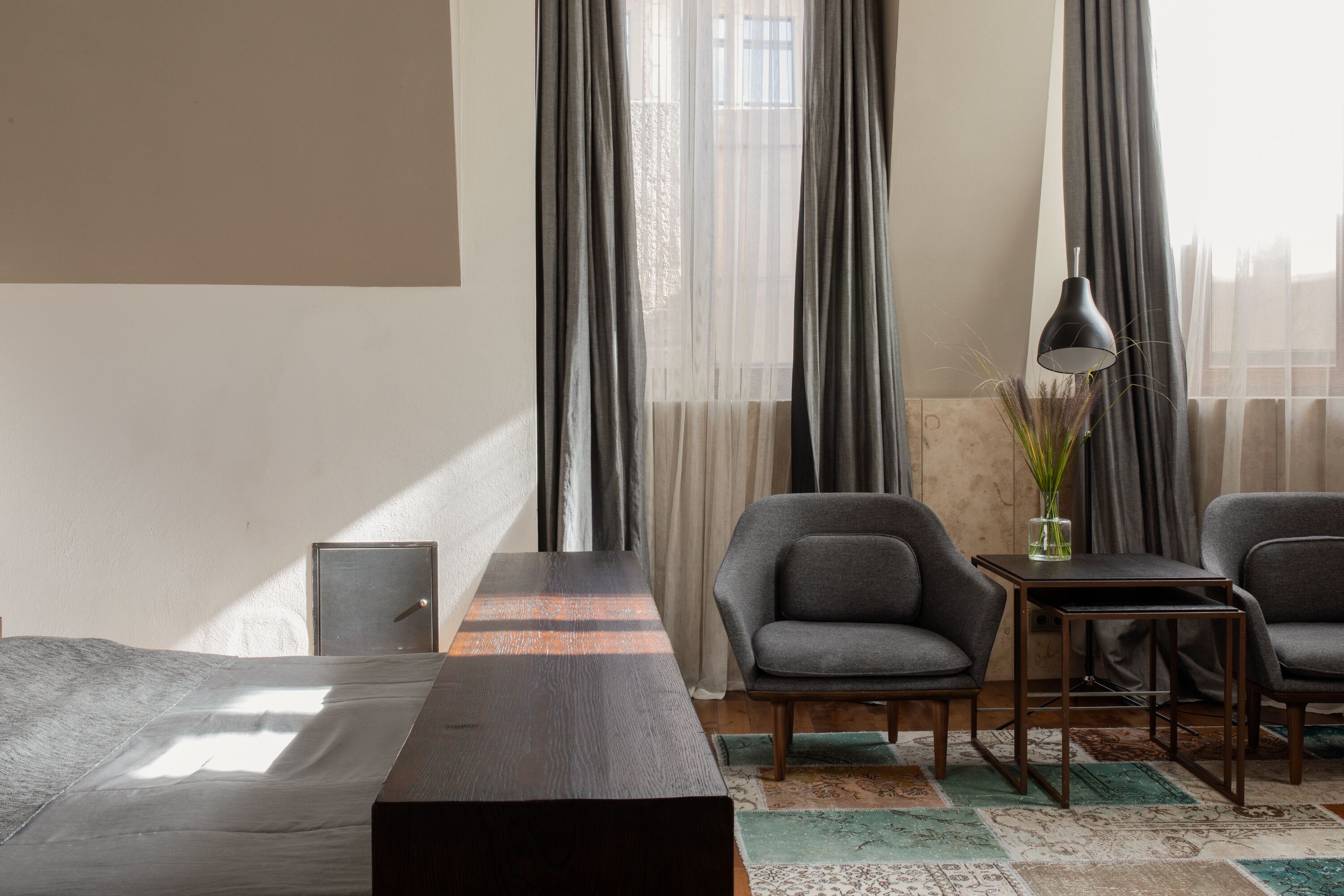 A hotel room with a bed, two armchairs, a small table, a lamp, and sheer curtains.