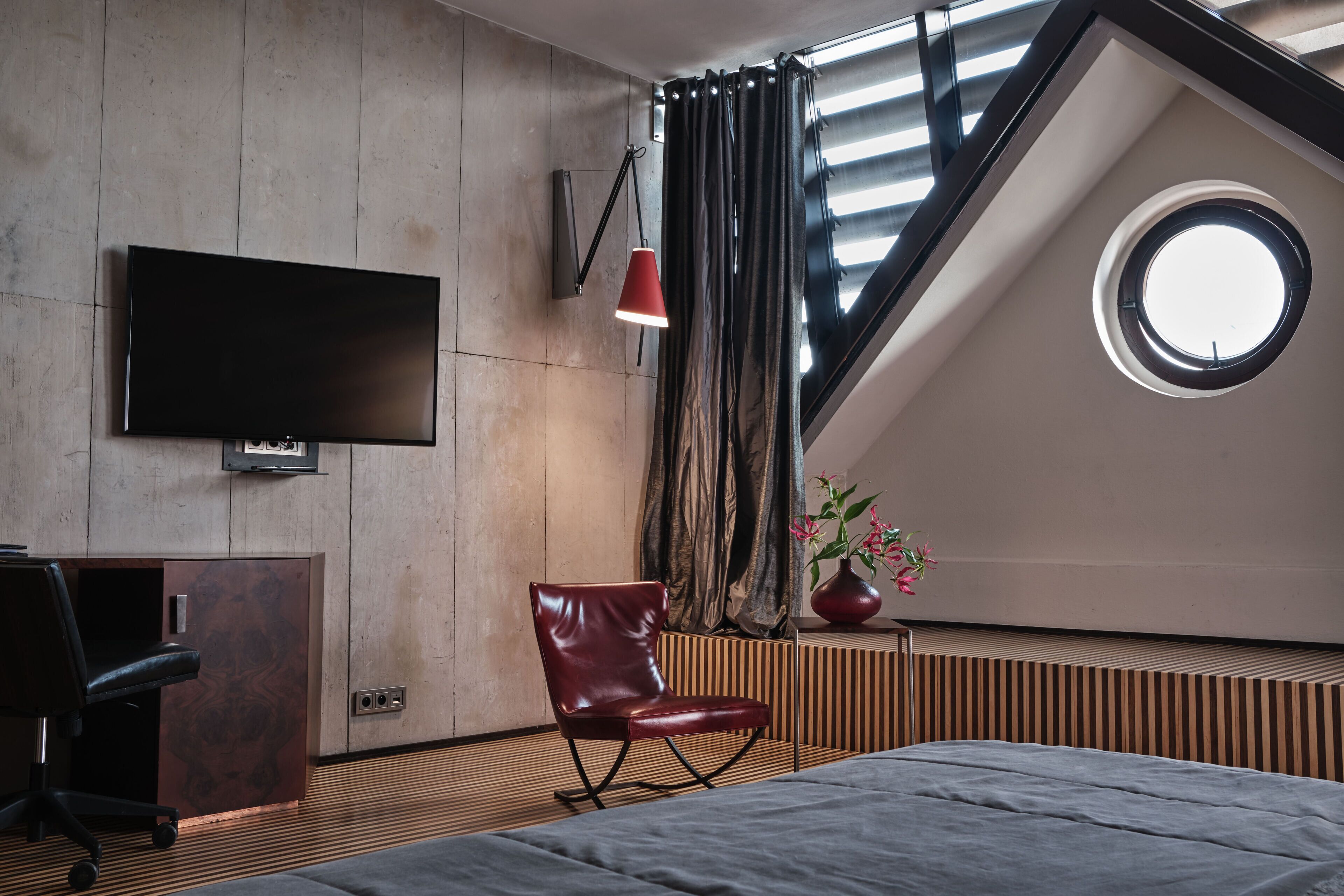 A modern bedroom with a large bed, a TV mounted on the wall, a red pendant light, a chair, and a window with curtains.