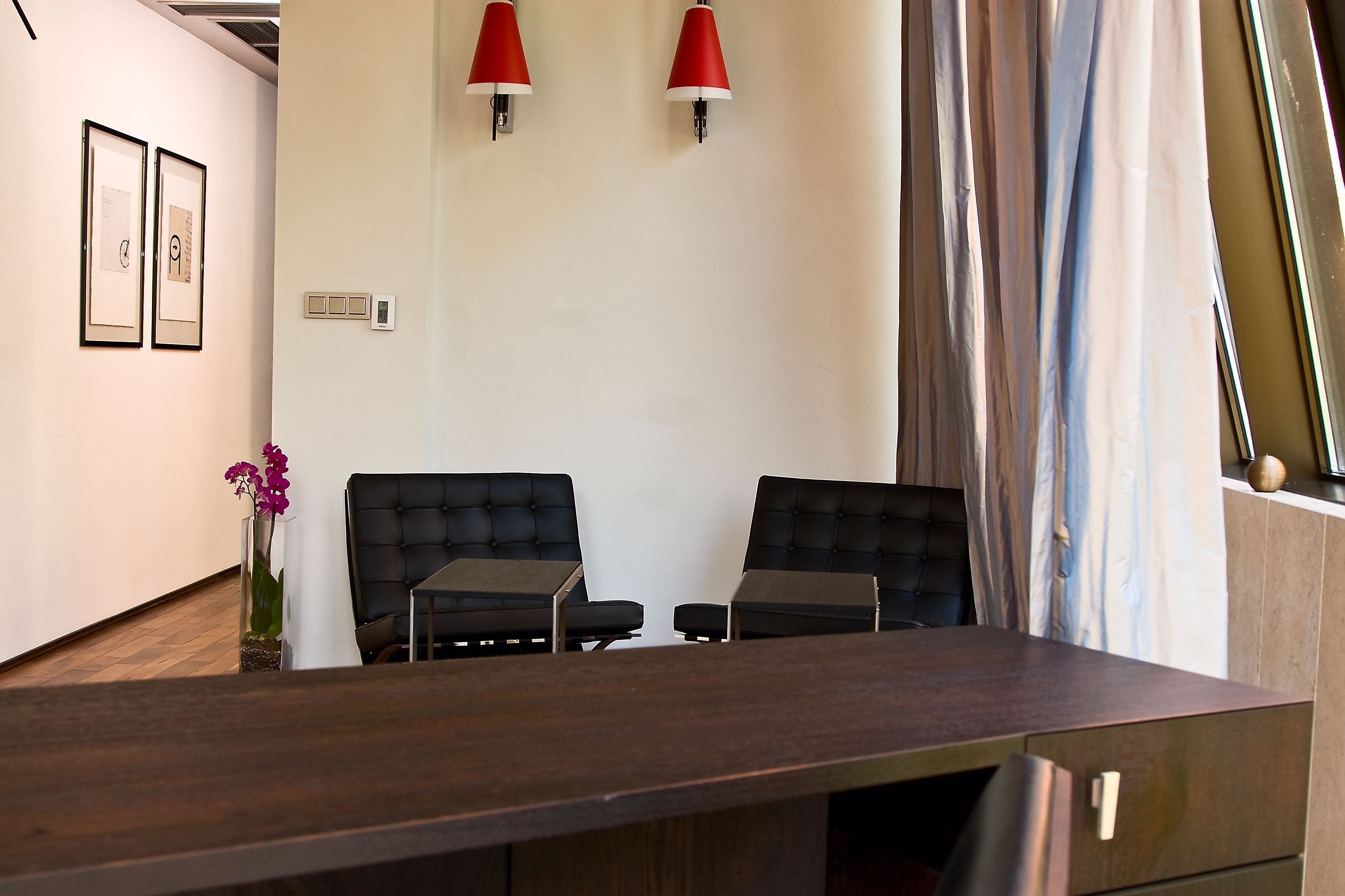 A modern office space with two black upholstered chairs, a wooden desk, and red wall-mounted lamps.