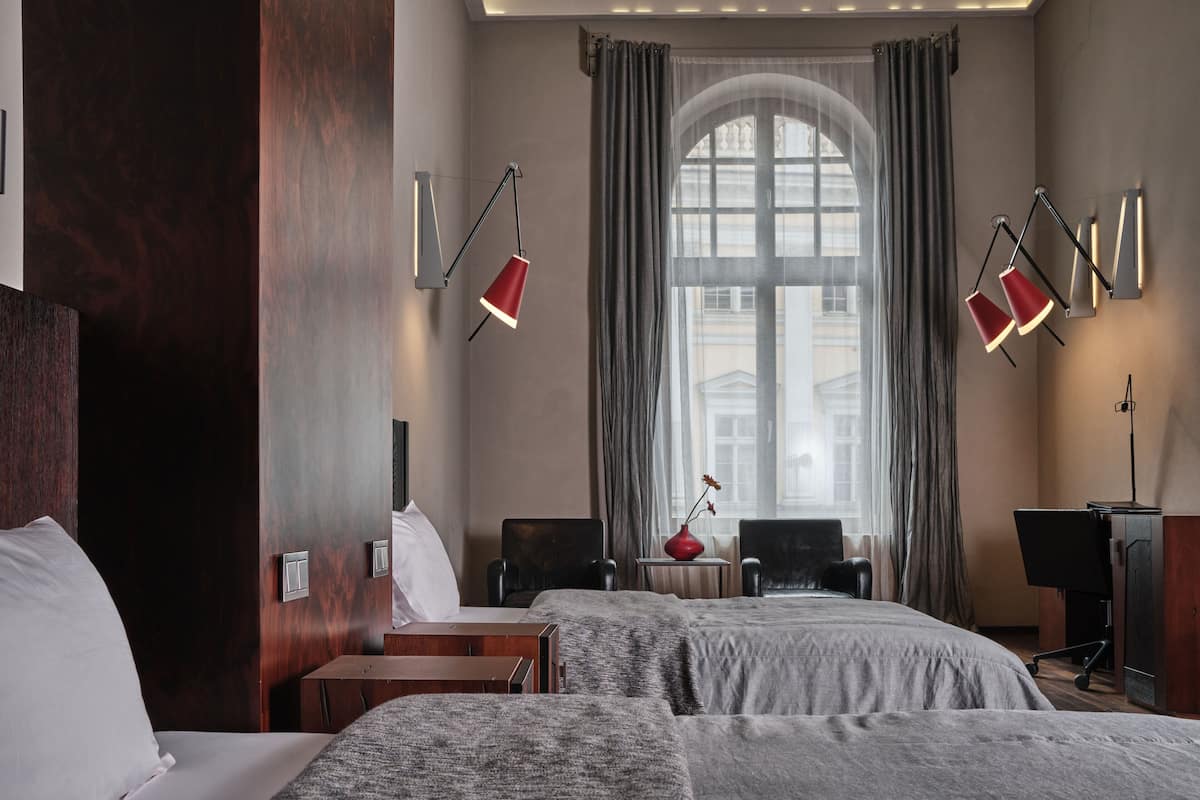 A hotel room with two beds, a desk, a chair, a small table, and a large window with curtains.