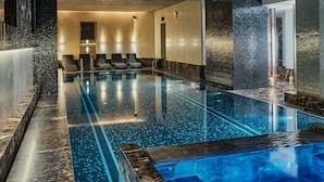 Una piscina interior moderna con una fuente de agua azul cristalina, un área de descanso con sillas y un ambiente tipo spa con iluminación cálida.