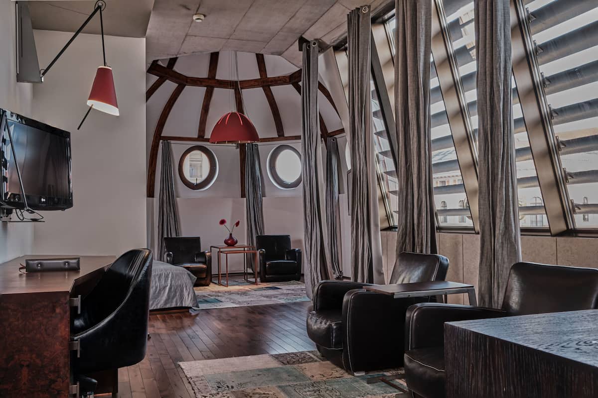 A modern hotel room with a wooden ceiling, a large TV, a dining area with a table and chairs, and large windows with curtains.