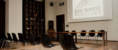 A conference room with a projector screen displaying "Hotel Monopol Wroclaw" logo, a wooden table, and black chairs arranged in rows.