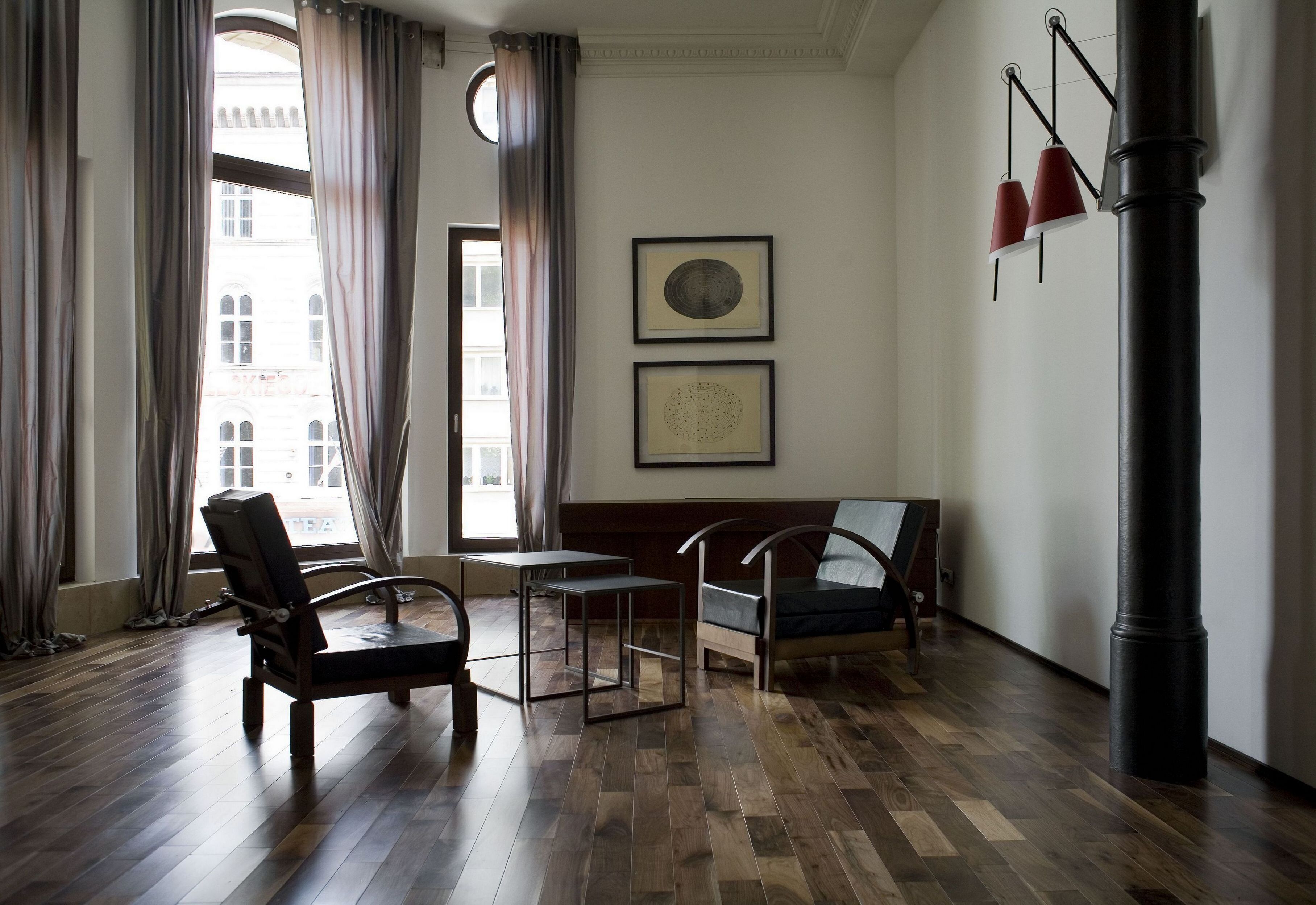 A room with wooden flooring, two chairs, a small table, a bench, a window with curtains, and two framed pictures on the wall.