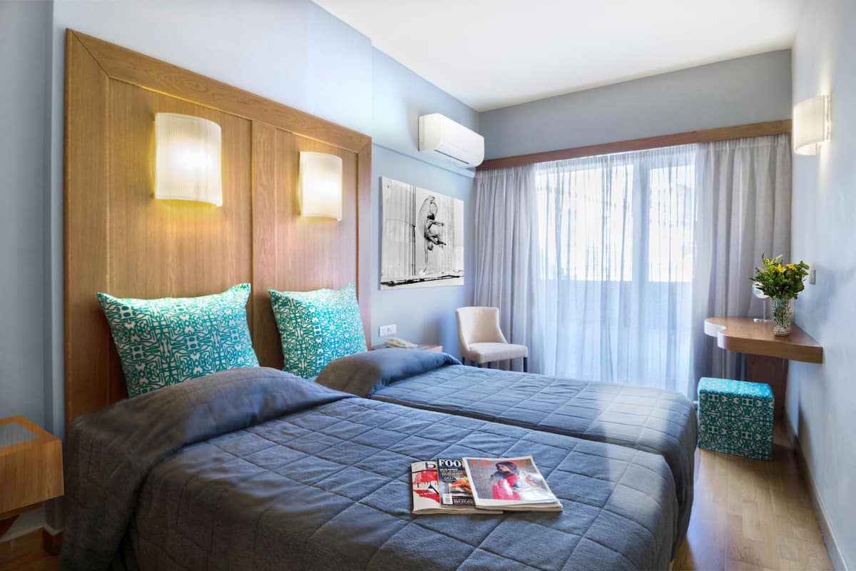 Une chambre d’hôtel avec deux lits, une tête de lit en bois, une chaise, une table de chevet, une petite table, une fenêtre avec des rideaux et un climatiseur fixé au mur.