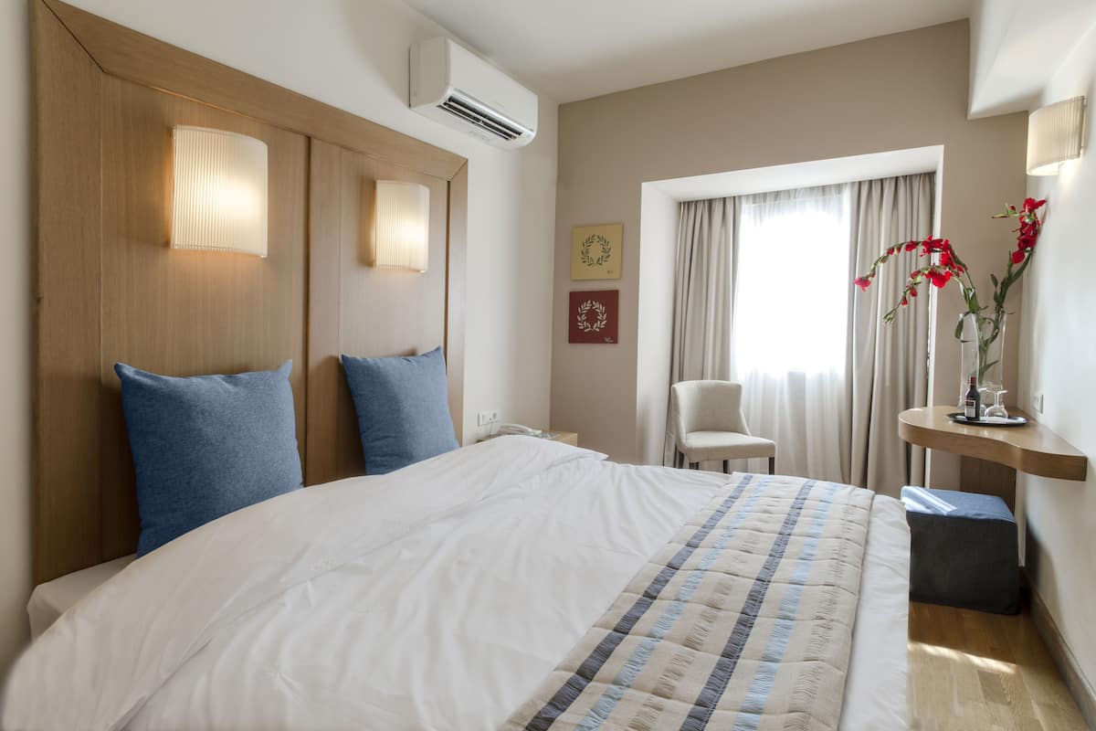 Une chambre d’hôtel avec un grand lit, deux lampes de chevet, une chaise, une petite table sur laquelle est posé un vase de fleurs et une fenêtre avec des rideaux.