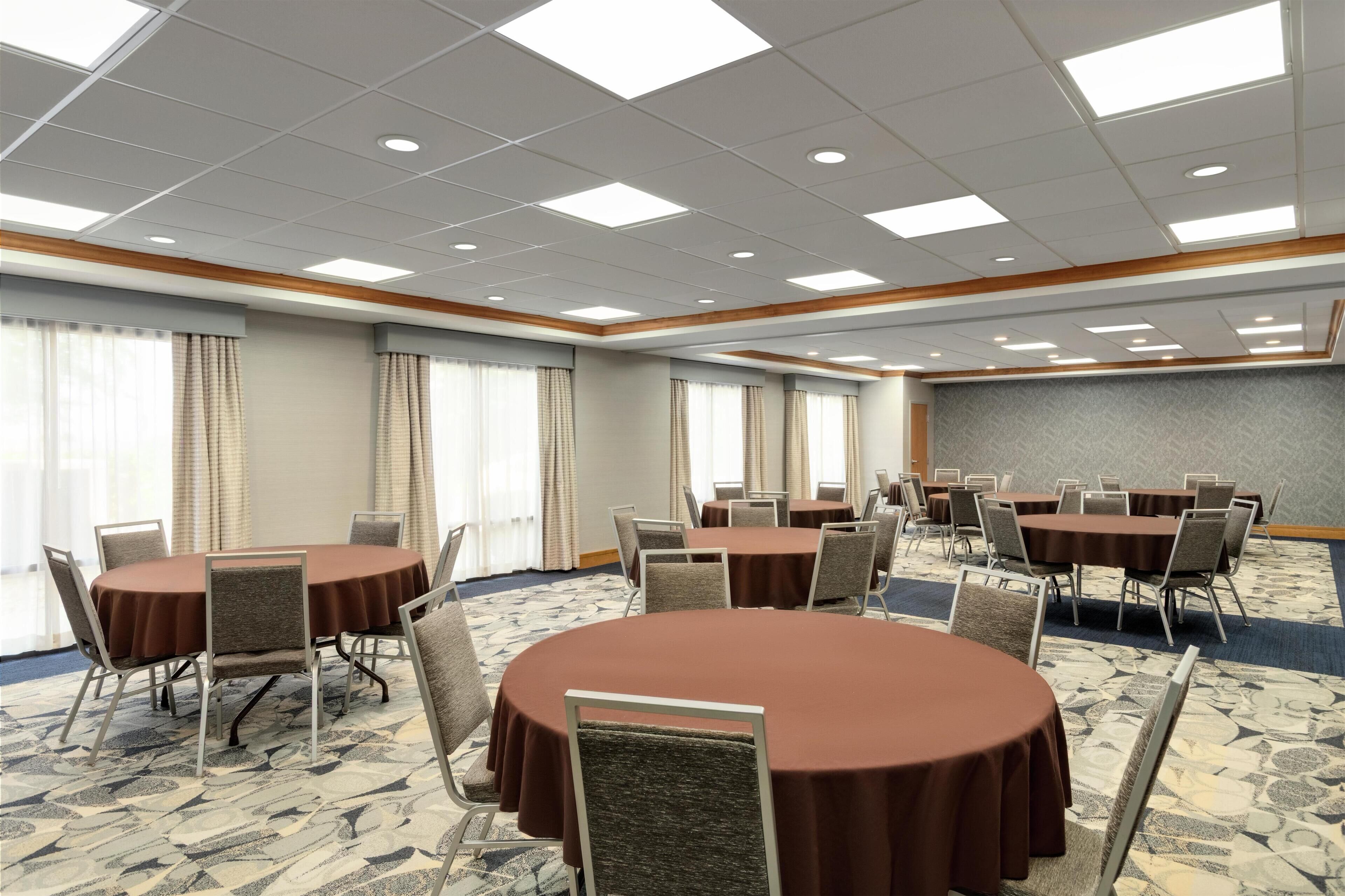 A conference room with multiple round tables and chairs, large windows with curtains, and a patterned carpet.