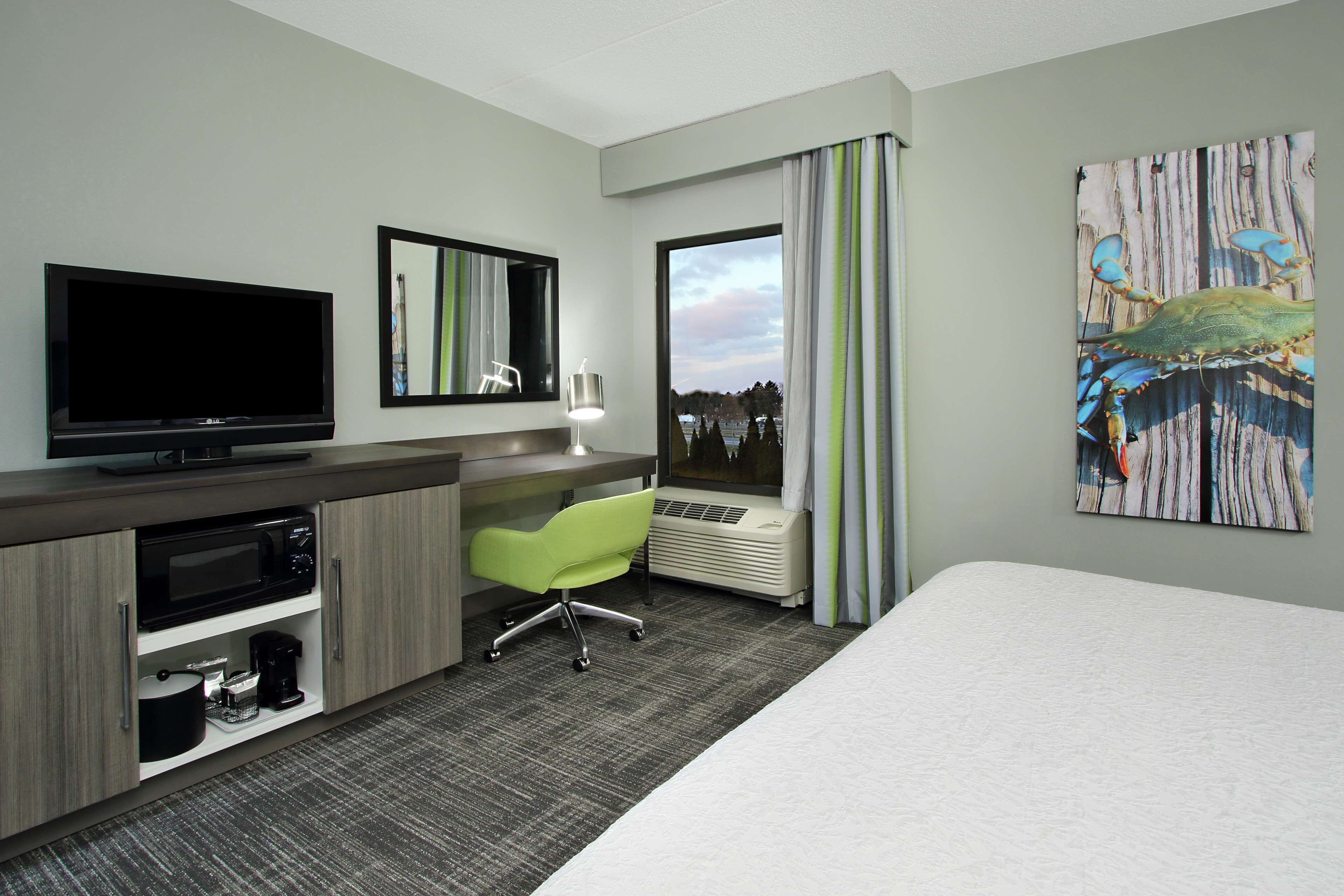 Habitación de hotel con cama, escritorio con televisión de pantalla plana, microondas, cafetera, silla verde, ventana grande con cortinas y un cuadro en la pared.